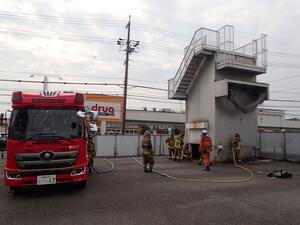 解体中の灰色の建造物の横に消防車両が配置され、複数人が訓練を行っている様子の写真