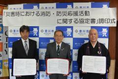 2人のスーツ姿の男性と1人の袈裟姿の男性が並んで、それぞれが協定書を両手で広げて立っている様子の写真