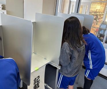 実際に選挙で使う記載台を使って、児童が投票用紙に候補者名を記入する様子の写真です。