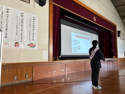 中部小学校の体育館で、市職員が模擬投票の候補者演説をする様子の写真です。