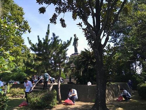 公園内のに生える多くの木と平和の像を清掃する人々の写真