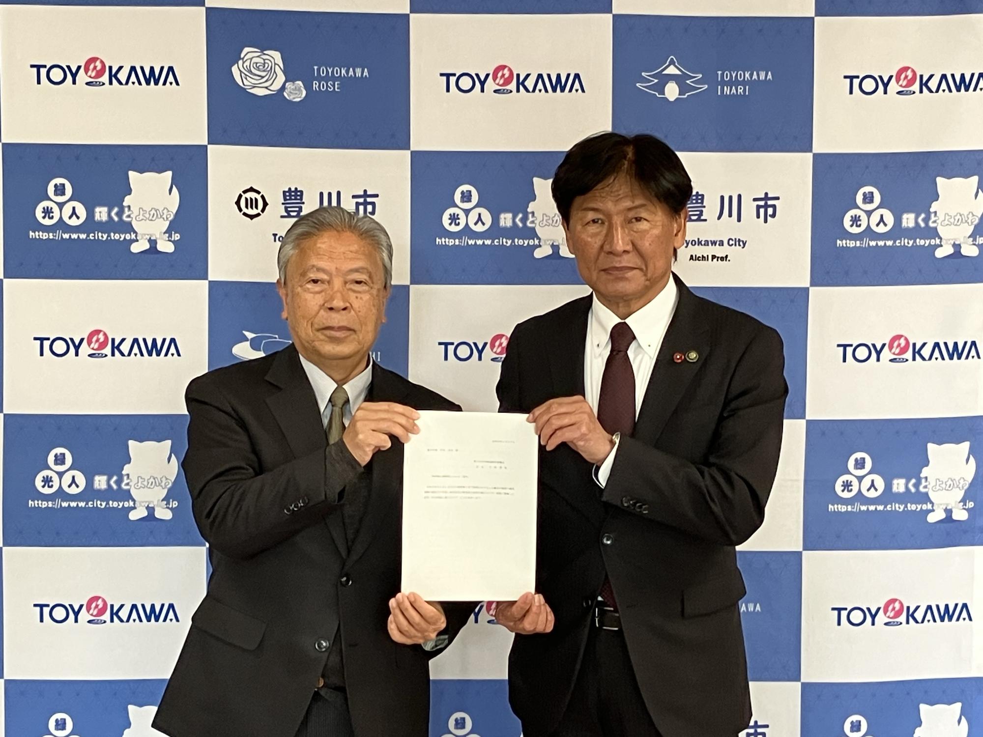 豊川市のバックパネルの前で、市長と特別職報酬等審議会会長が答申書を持ち、こちらに顔を向けている写真