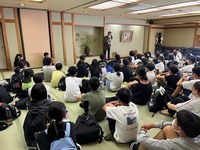旅館の宴会場で、畳に座りがなら教職員の話を聞く大勢の6年生の写真