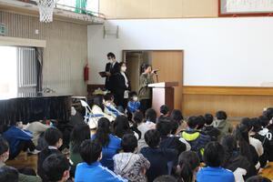 児童が体育館で児童集会の司会を行い体育座りの児童たちに話をしている写真