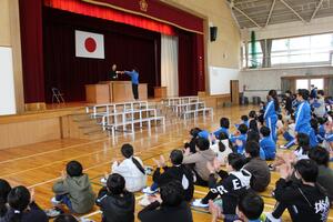 児童と教諭が体育館のステージ台で表彰披露を行い他の児童たちが体育座りで拍手をしている写真