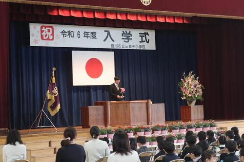 「祝 令和6年度 入学式」と書かれた弾幕が掲げられたステージに男性が立っている写真