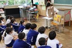 教室で大勢の児童たちが体育座りをして座っており、その前で椅子に座った大人が本を開いている写真