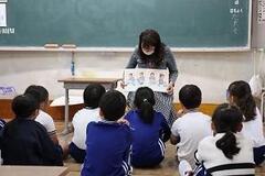 教室で大勢の児童たちが体育座りをして座っており、その前で椅子に座った大人がイラストが付いた用紙を児童側に見せている写真