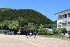 空の青と山の緑を背景にした校庭で、白線のレーンを走る児童達の写真
