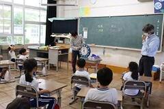 教室内で席に着席する児童達の前で、大きな時計の文字盤を手に持ち指差す教師の写真