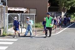ランドセルを背負い道路の端から横断歩道へ歩く児童達と、それを見守る帽子を被り緑の服を着た男性の写真