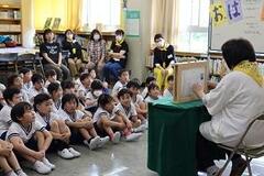 室内で大勢の児童たちが体育座りをしており、その前で大人が椅子に座っている写真
