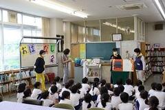 室内で大勢の児童たちが体育座りをしており、その前で大人が立ったまま本を見開いている写真