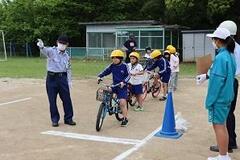 黄色いヘルメットをかぶった児童たちが白い線の前で列になって自転車に乗って並んでおり、一人の大人が遠くのほうを指さしている写真