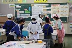 室内で給食服に身を包んだ児童たちが机の上で作業をしている写真