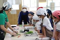 室内の机の上で大人が包丁を持ち、側には野菜が置かれ、児童たちがそれを見つめている写真