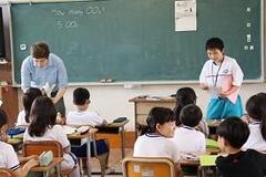教室の黒板の前に2名の大人が立っており、席に座っている複数の児童がそちらを見ている写真