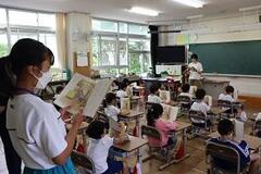 教室の後方で立って教科書を読んでいる児童と、席について聞いている児童たちの写真