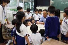 教室で椅子に座った一人の大人が作業している手元を、周りに集まった児童たちが眺めている写真