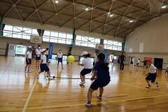体育館で黄色いボールが飛んでおり、その周りで児童が立っている写真