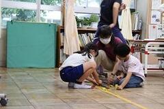 教室の床にしゃがみ作業をしている2名の児童とそれを見ているひとりの大人の写真