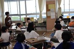 教室の窓を背景に、一人の児童が立ち上がっていて、大勢の児童たちが席に座っている教室の写真
